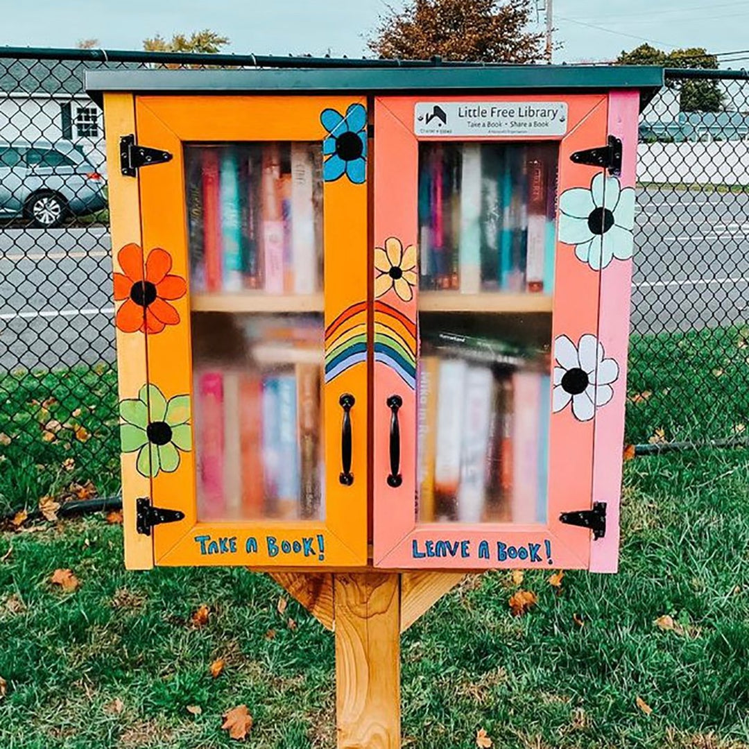 A little free library built on a grassy lawn. The sides are painted orange and pink with flowers around it. Inside, it is full of books.